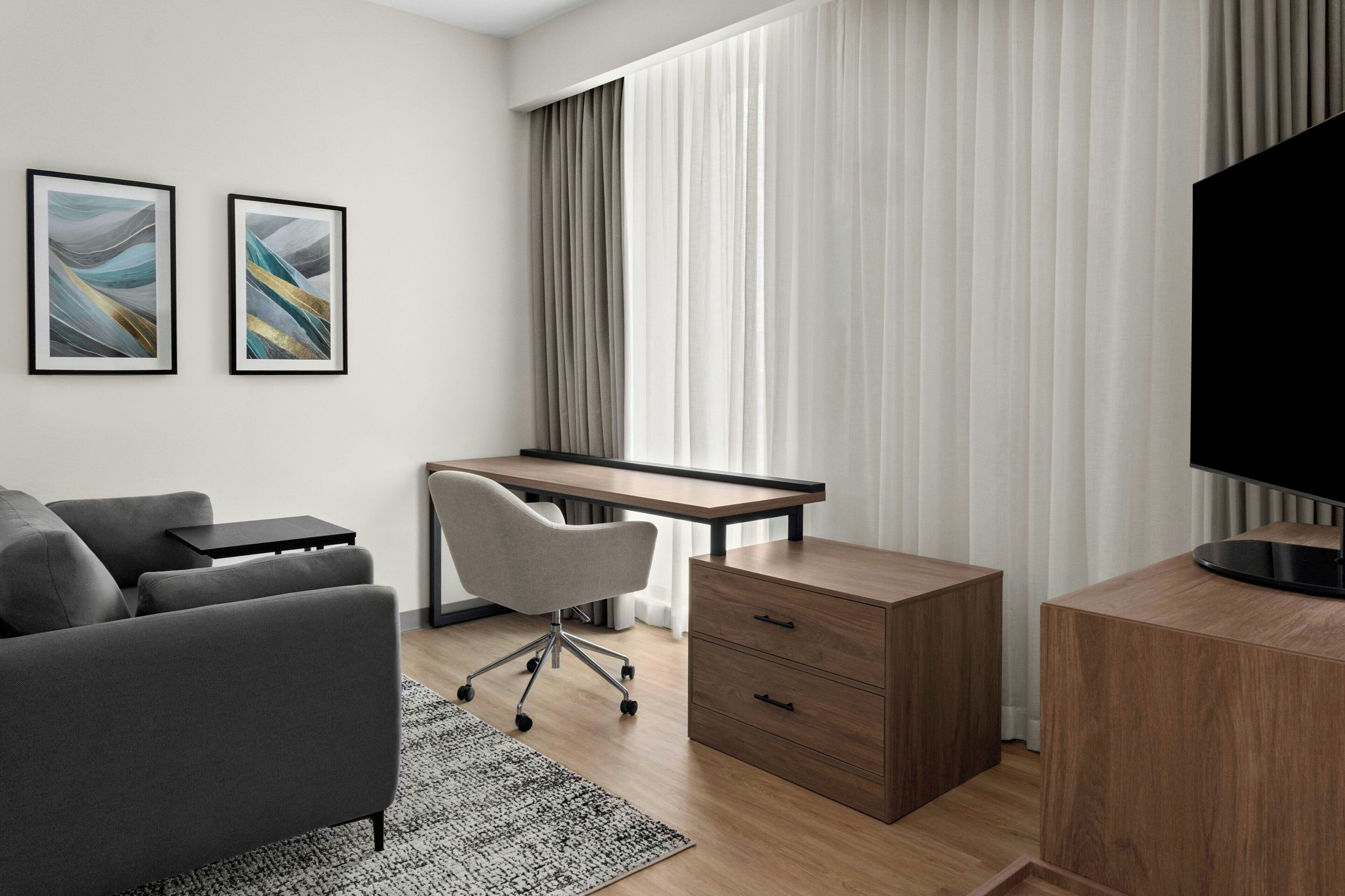 Moderni hotellihuone, jossa on sohva, työpöytä, tuoli, televisio ja taideteos seinällä.