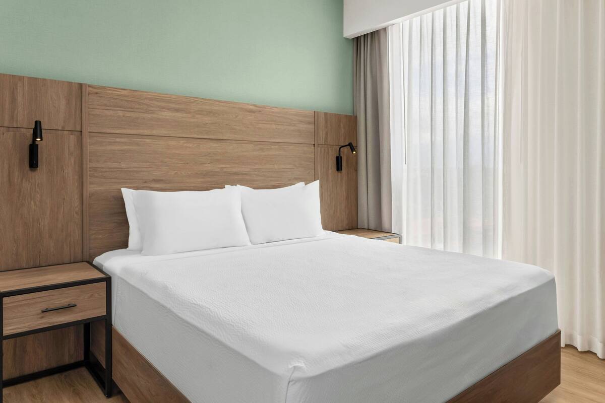 Una cama bien hecha con cabecera de madera, una mesita de noche con cajón y una ventana con cortinas traslúcidas.