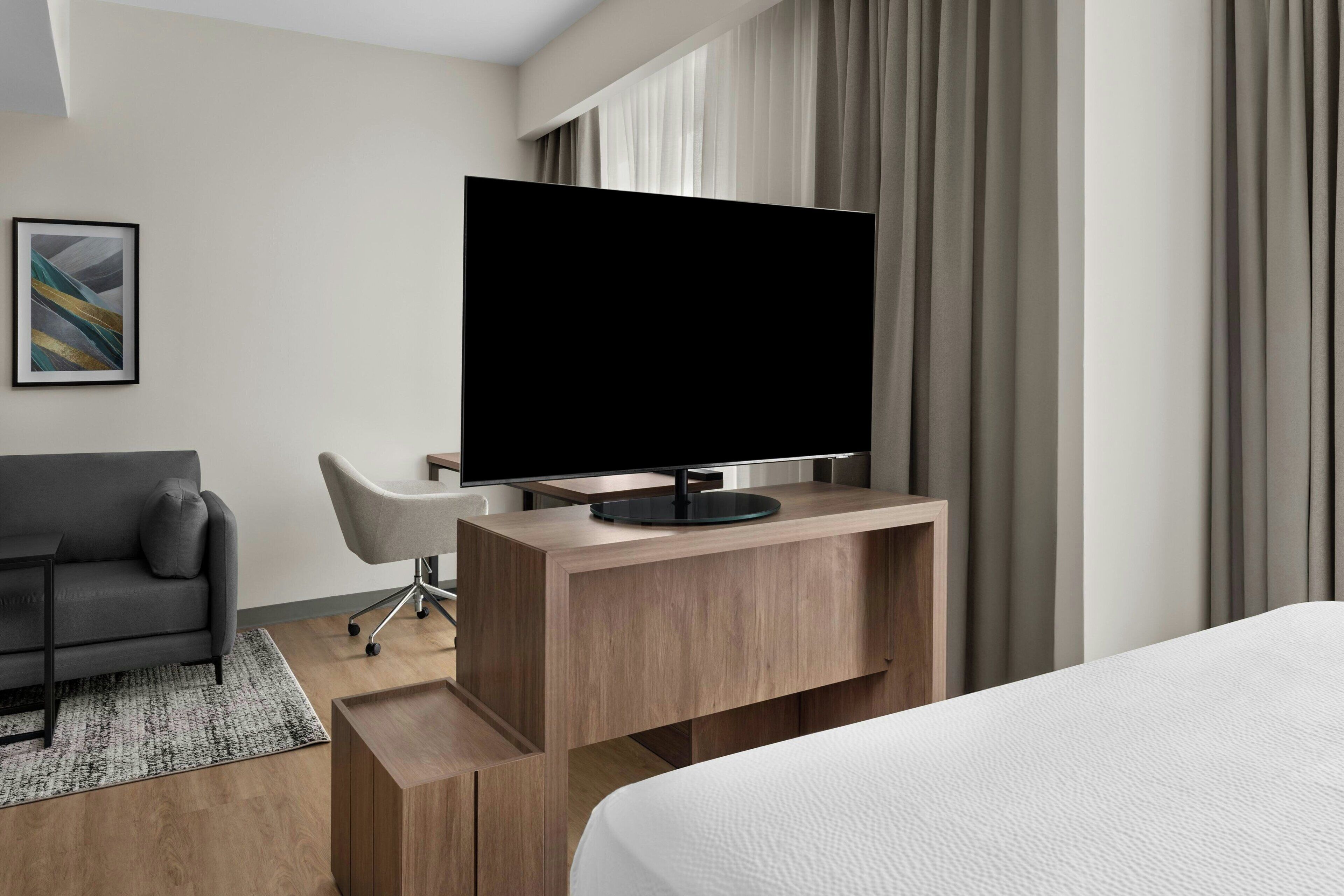A modern hotel room with a large bed, a flat-screen TV on a wooden console, a grey sofa, and a desk with a chair.