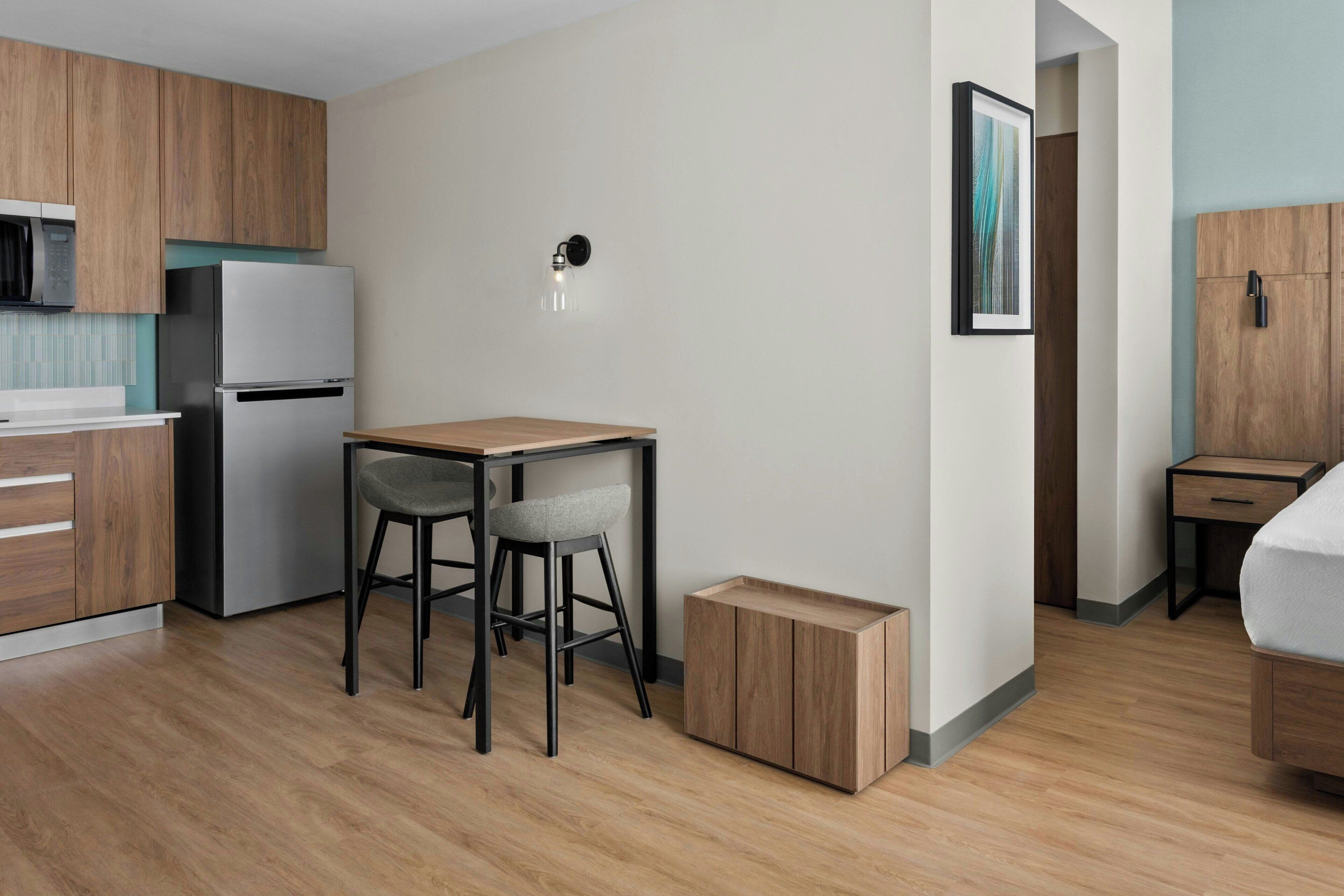 Una cocina compacta con refrigerador, una pequeña mesa de comedor con dos sillas, una lámpara y un mueble de madera.