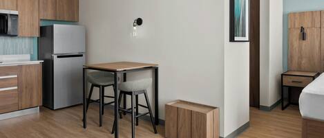 Una cocina compacta con refrigerador, una pequeña mesa de comedor con dos sillas, una lámpara y un mueble de madera.