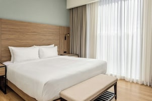 A modern hotel room with a wooden headboard, a large bed with white linens, a bedside table, a bench at the foot of the bed, and sheer curtains.