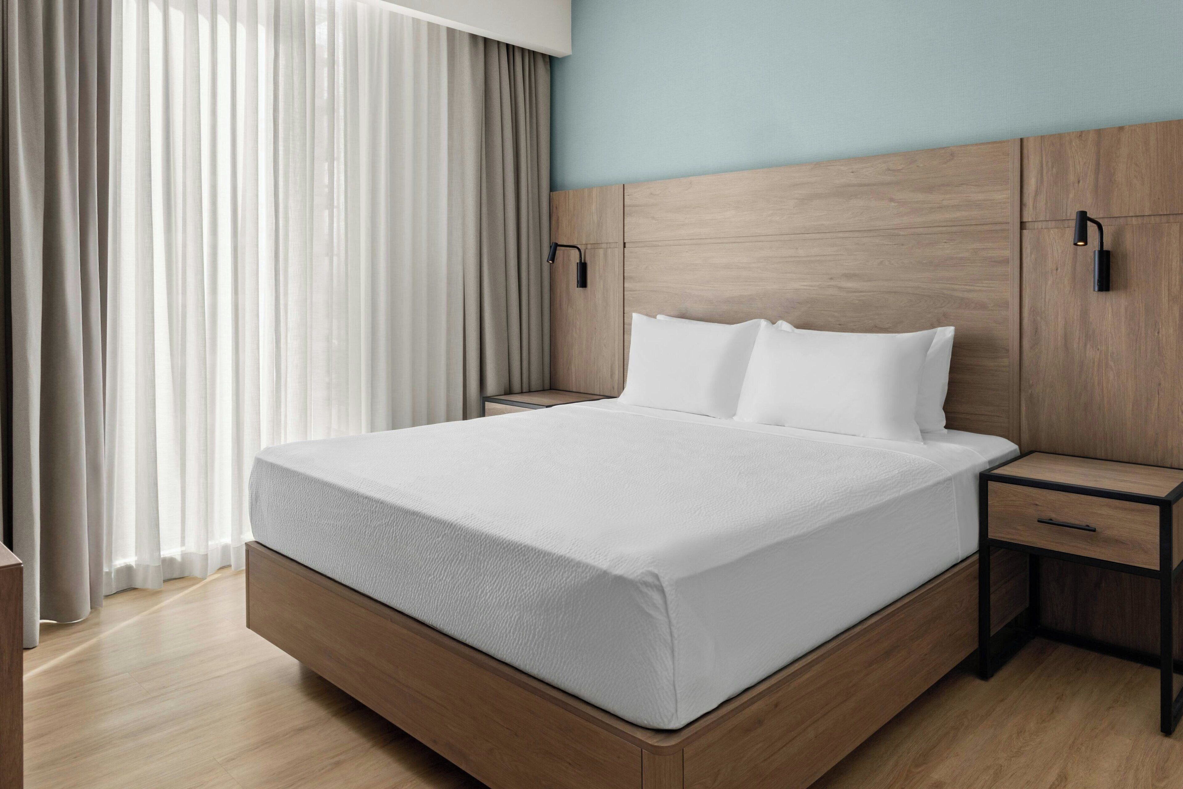 Una habitación de hotel moderna con cabecera de madera, una cama grande con ropa blanca y una mesita de noche con cajón, además de cortinas translúcidas.
