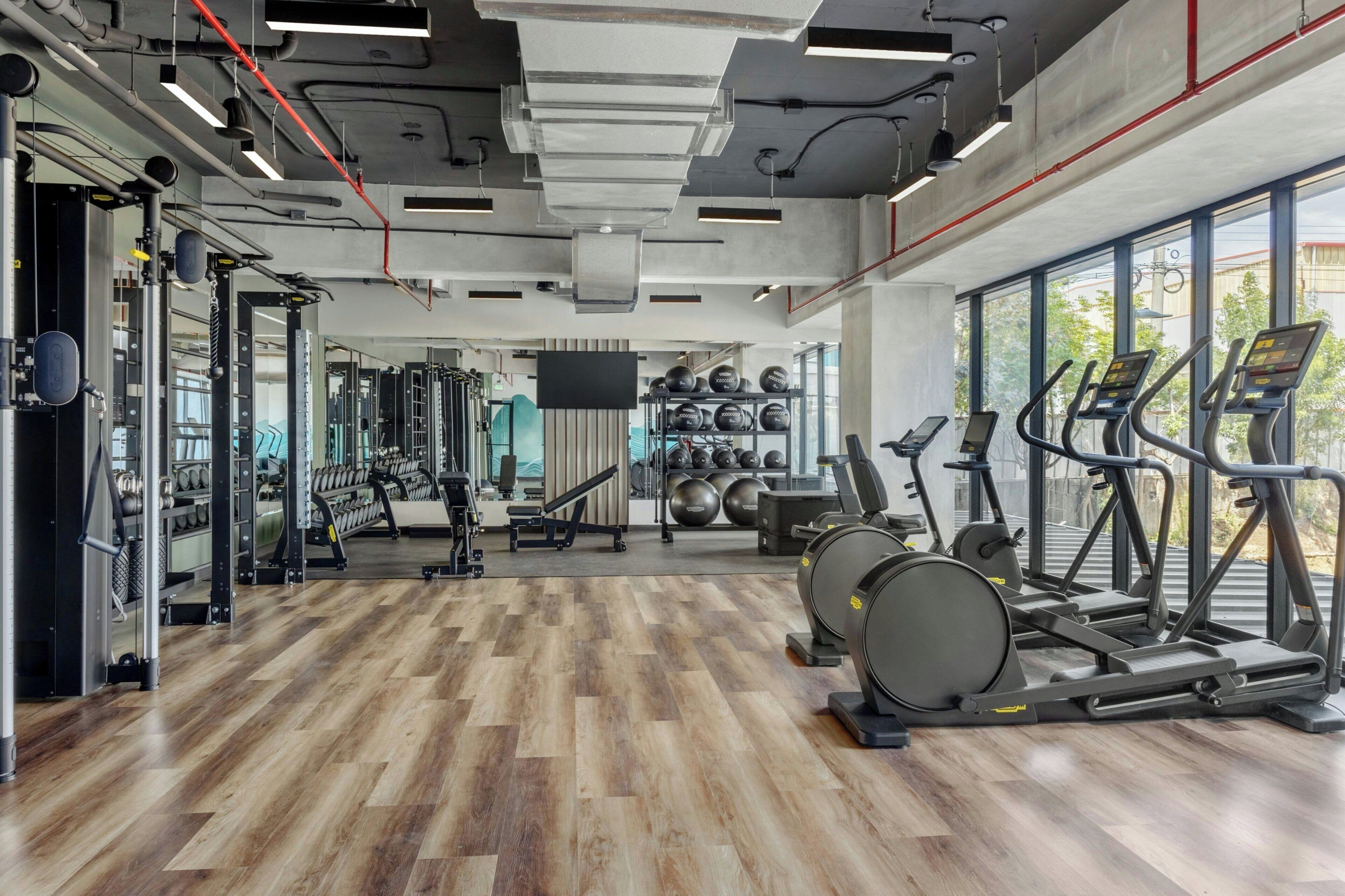 Un gimnasio con equipamiento de ejercicio, piso de madera y ventanas amplias.