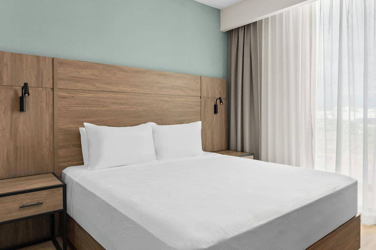 Una habitación de hotel moderna con cabecera de madera, una cama grande con ropa blanca, una mesita de noche con cajón y una ventana con cortinas traslúcidas.