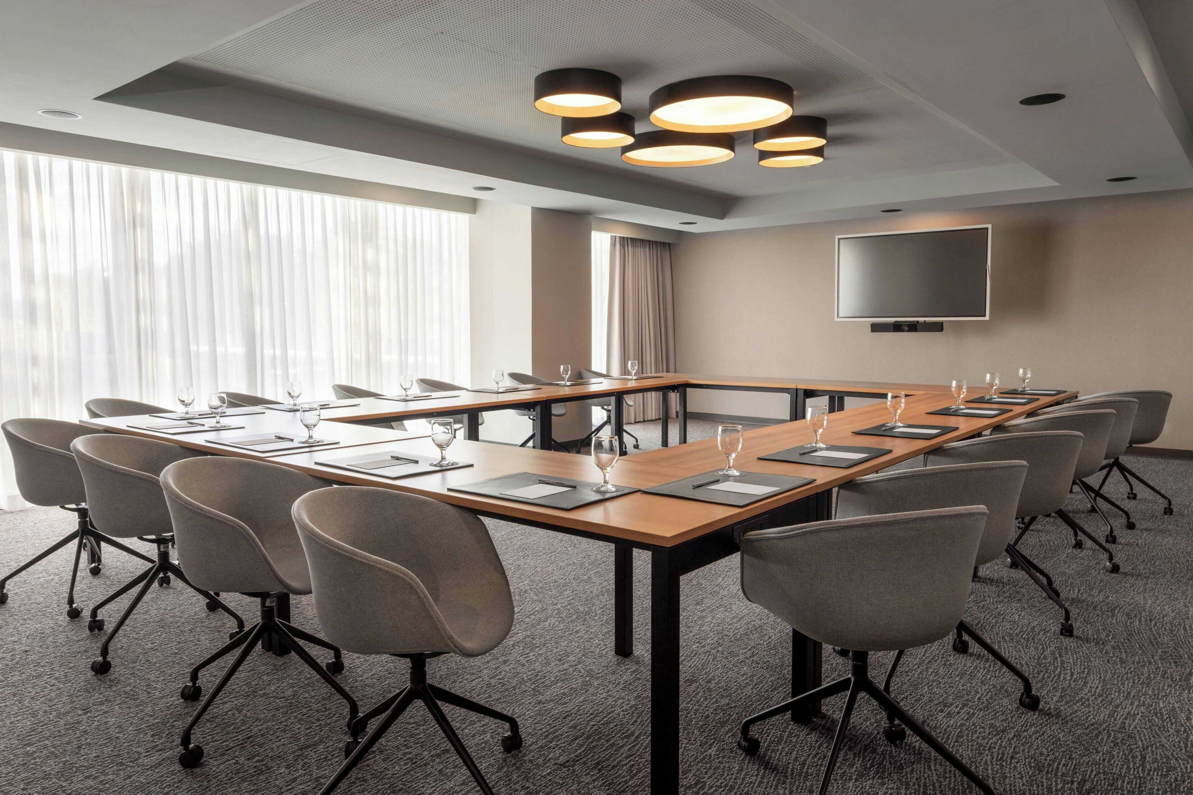 A conference room with a long table, chairs, a flat-screen TV, and a large window with curtains.