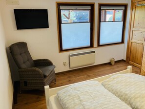A room with a bed, a chair, a TV, two windows with blinds, a heater, and a wooden wardrobe.