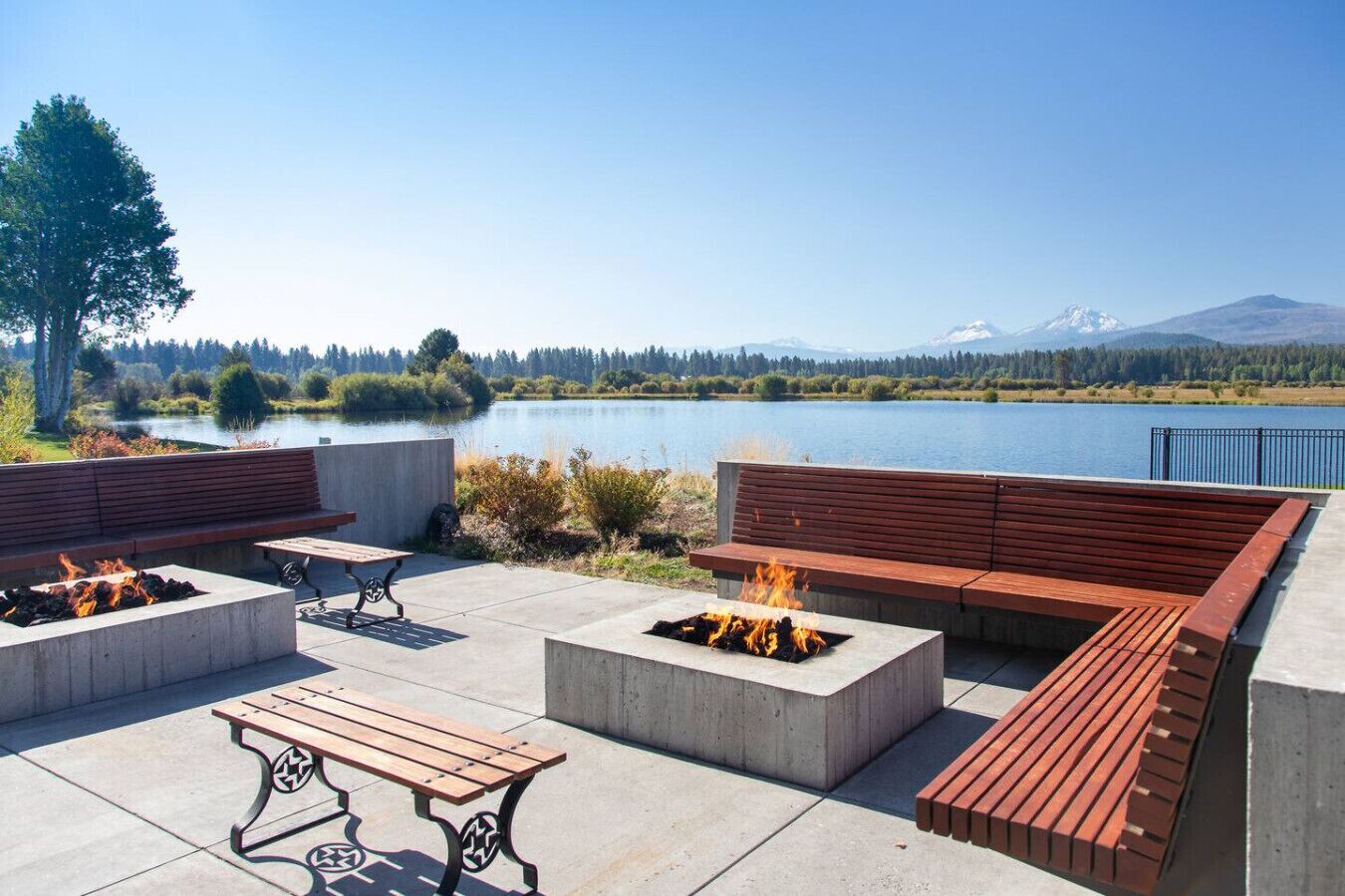 An outdoor seating area with concrete planters, wooden benches, and a fire feature, overlooking a lake and distant mountains.