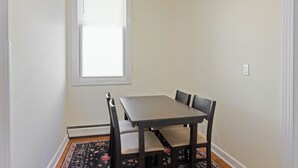 A room with a dining table and four chairs, a window with blinds, and a patterned rug on the floor. - All-American classic/ 18 min to Manhattan (Carlstadt)