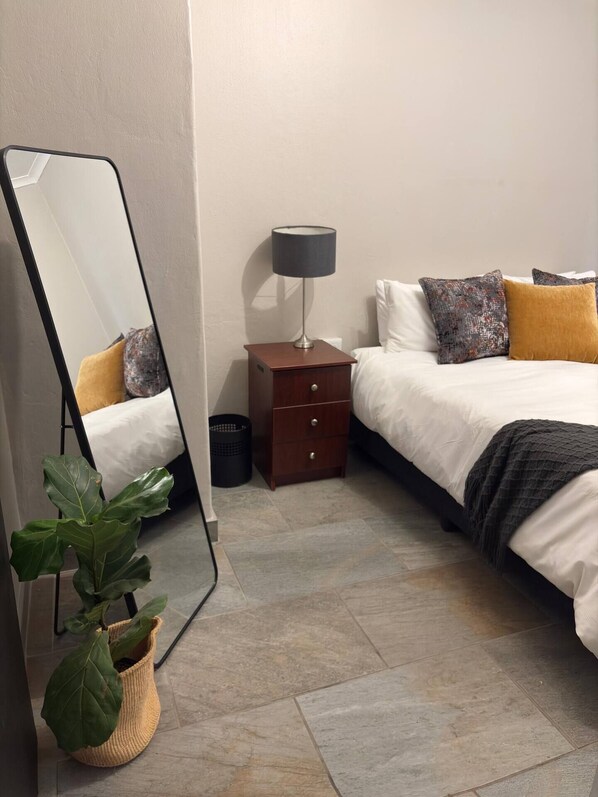 A neatly arranged bedroom with a large mirror, a bedside table with a lamp, a bed with pillows, and a potted plant.