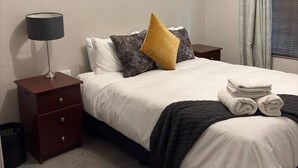 Ein ordentlich bezogenes Bett mit weißen Bettwäsche, einem senfgelben Kissen und einer schwarzen Tagesdecke. Ein kleiner Holfnachtschränkchen mit Lampe und zusammengelegtem Handtuch. Der Raum hat ein schlichtes, sauberes Design mit Fliesenboden und einem Fenster mit Vorhängen.