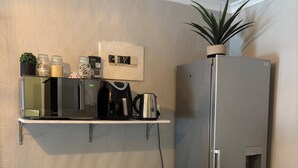 Eine Küche mit Mikrowelle, Kaffeemaschine und Wasserkocher auf einem Regal, einem Kühlschrank, einer Mülltonne und einem Holztisch.