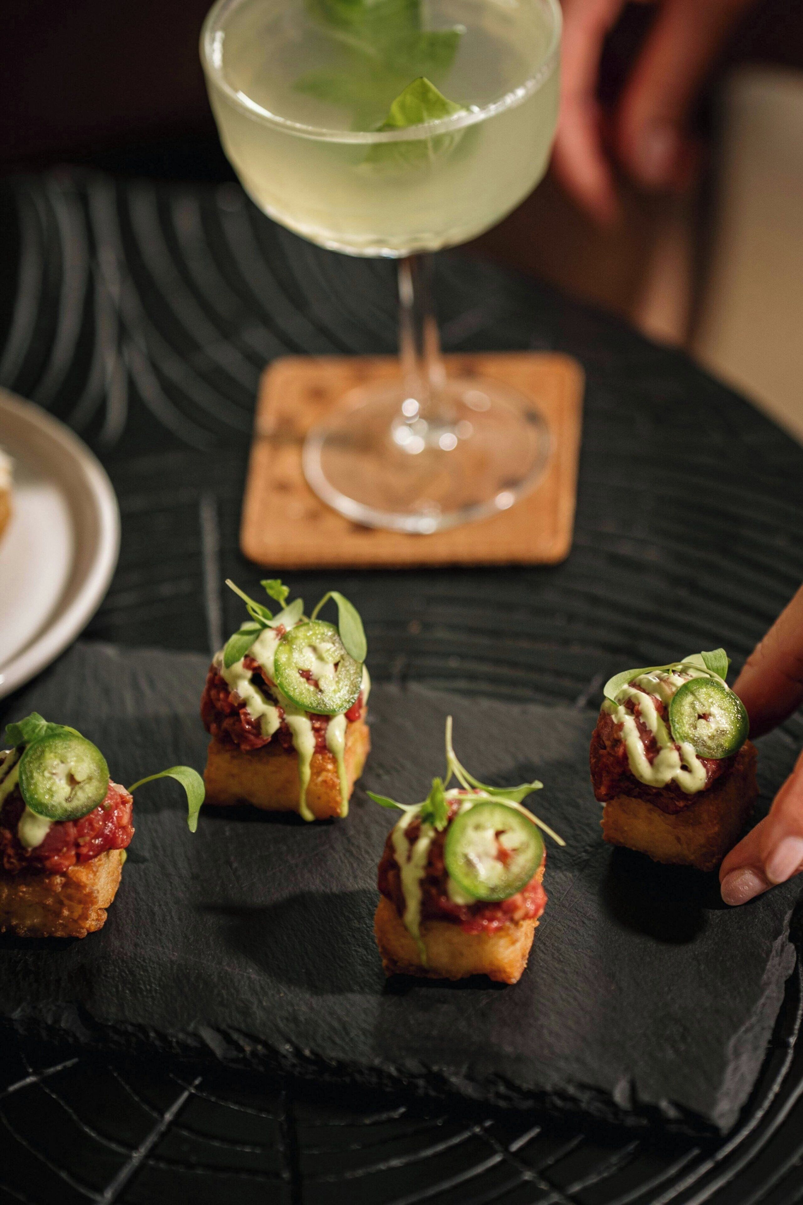 Une table avec un plateau en ardoise noire et quatre petites bouchées garnies de rondelles de piment jalapeño et d’une sauce blanche, accompagnées d’un verre à cocktail décoré d’une feuille de menthe.