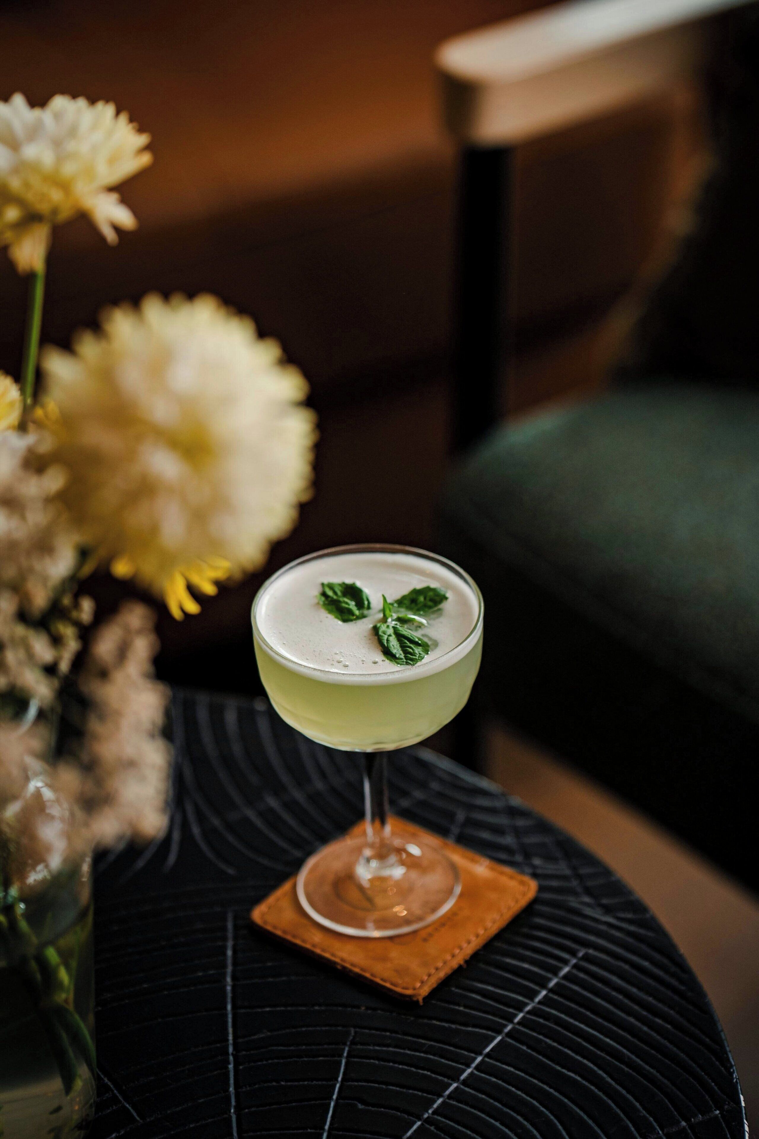 A cocktail in a glass on a coaster, with a blurred background of flowers and furniture.