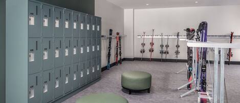 A locker room with multiple lockers, a ski rack, and two green ottomans.
