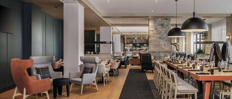 A modern hotel lobby with a long dining table, multiple seating areas, and a bar.