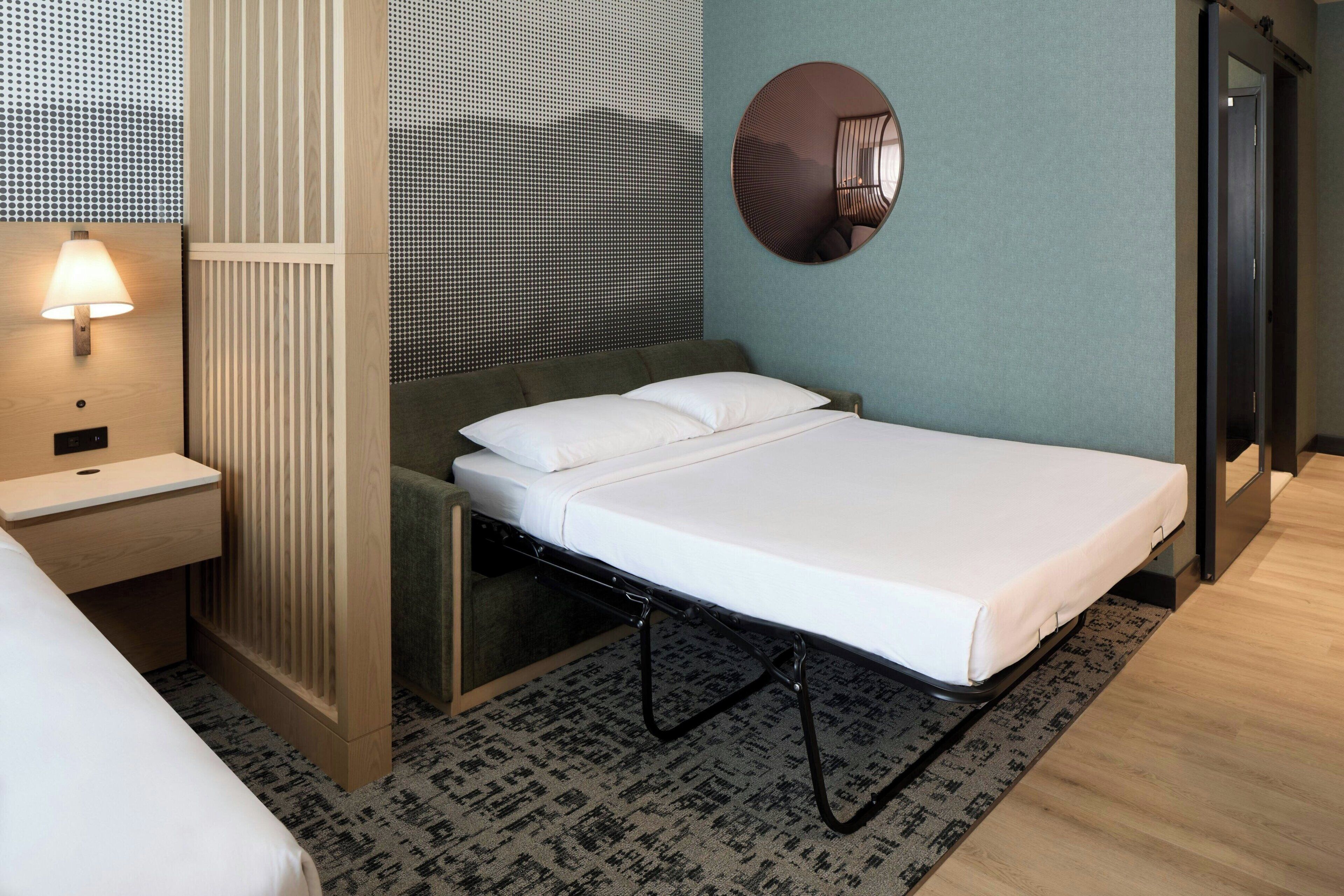 Una habitación de hotel moderna con una cama individual, una mesita de noche, una lámpara, un espejo y una alfombra estampada.