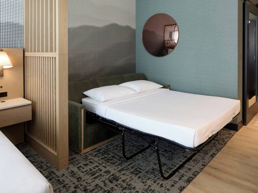 Una habitación de hotel moderna con una cama individual, una mesita de noche, una lámpara, un espejo y una alfombra estampada.