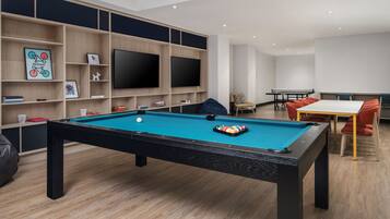 A pool table with a blue felt surface, surrounded by wooden shelves with various items, a flat-screen TV mounted on the wall, and a seating area with a table and chairs.