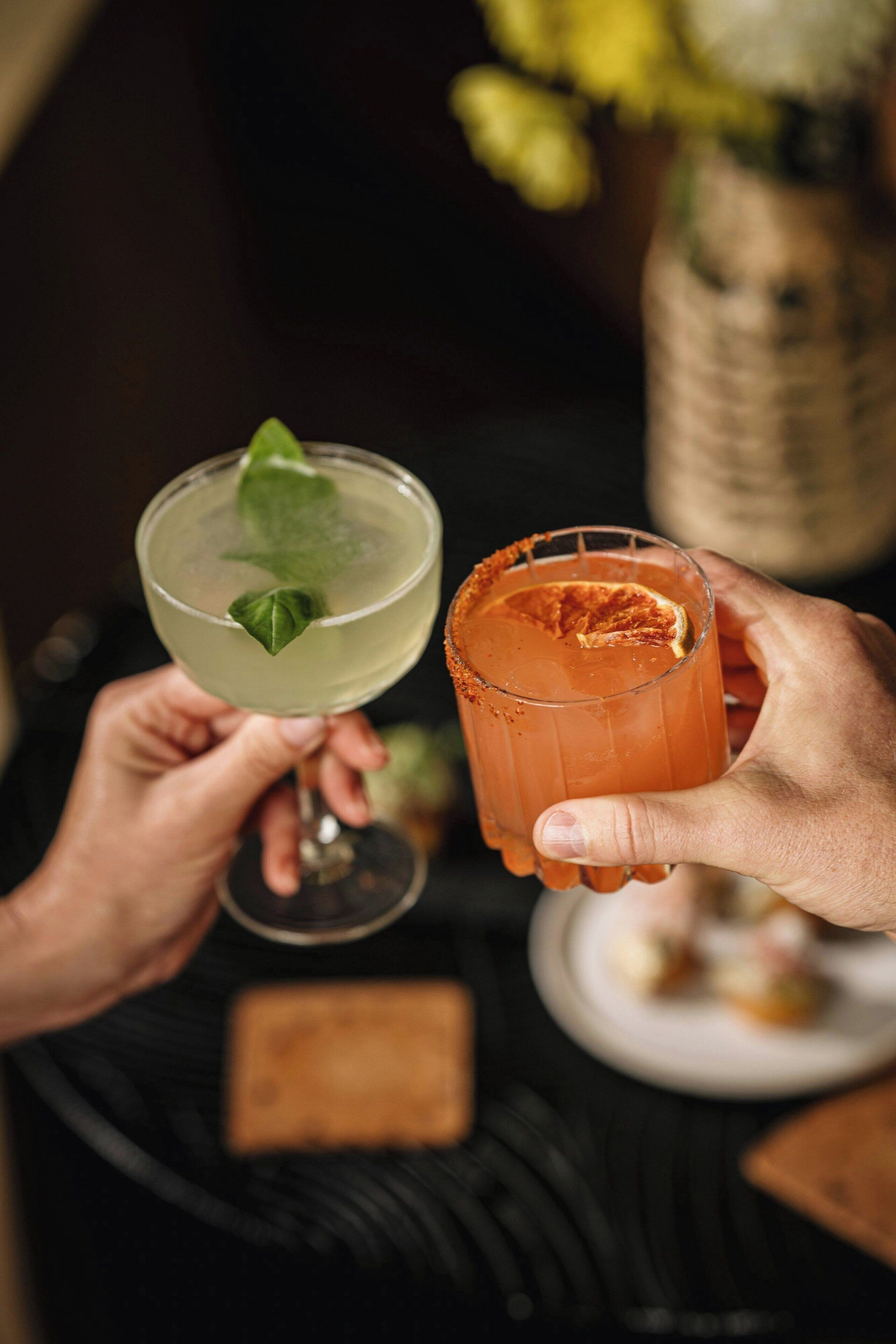 Two hands holding cocktail glasses with a blurred background.
