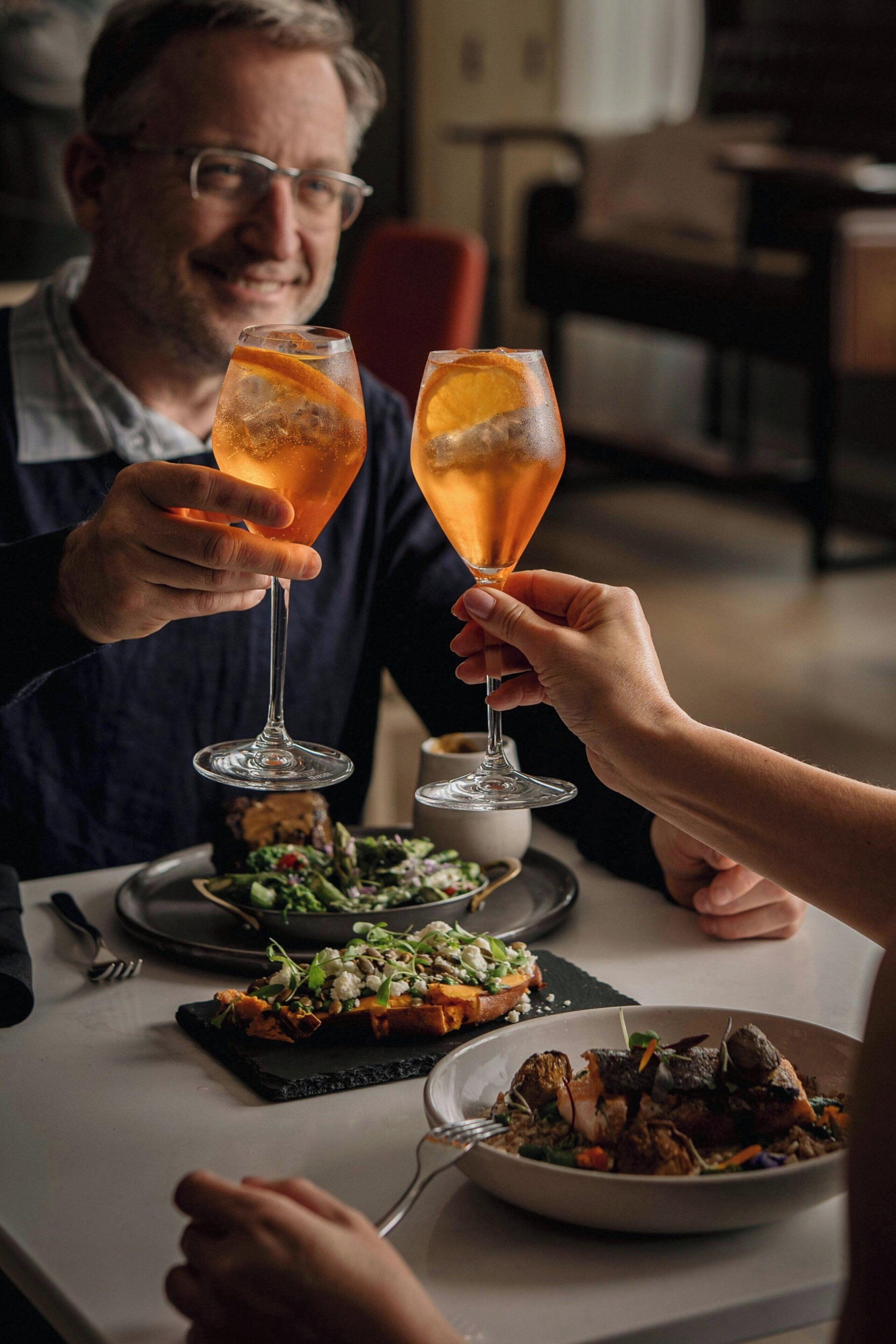 A dining table with two people toasting with glasses, a plate of food, and a bowl of salad.