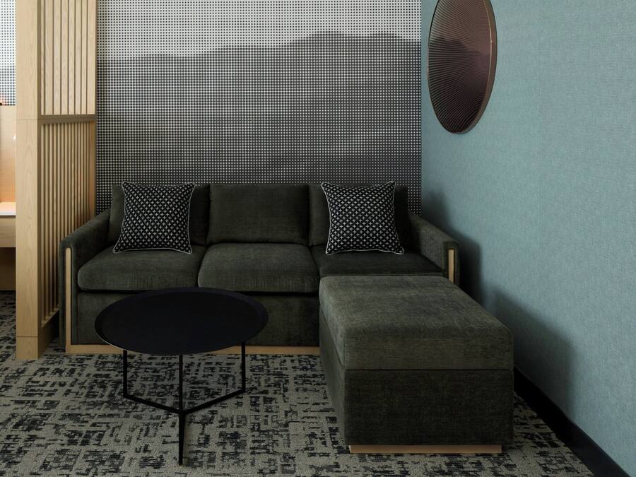 Una habitación de hotel moderna con un sofá gris, un reposapiés negro, una mesita pequeña negra y una alfombra estampada.