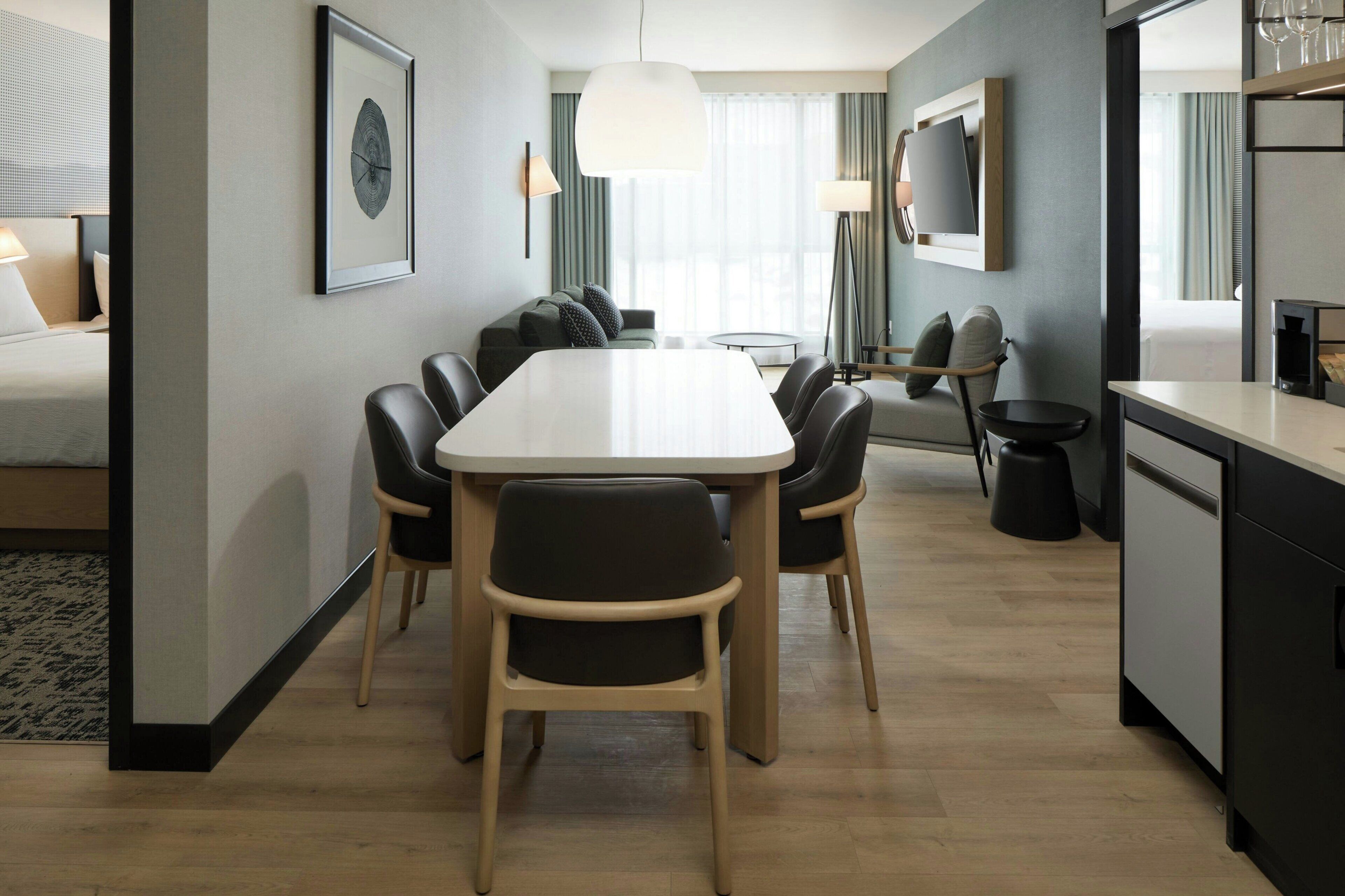 A modern hotel room with a dining area, a bed, a TV, and a kitchen area.