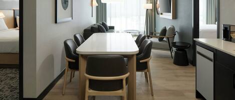 A modern hotel room with a dining area, a bed, a TV, and a kitchen area.
