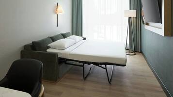 Una habitación de hotel moderna con sofá cama tipo futón, televisor de pantalla plana en la pared y una lámpara de pie.