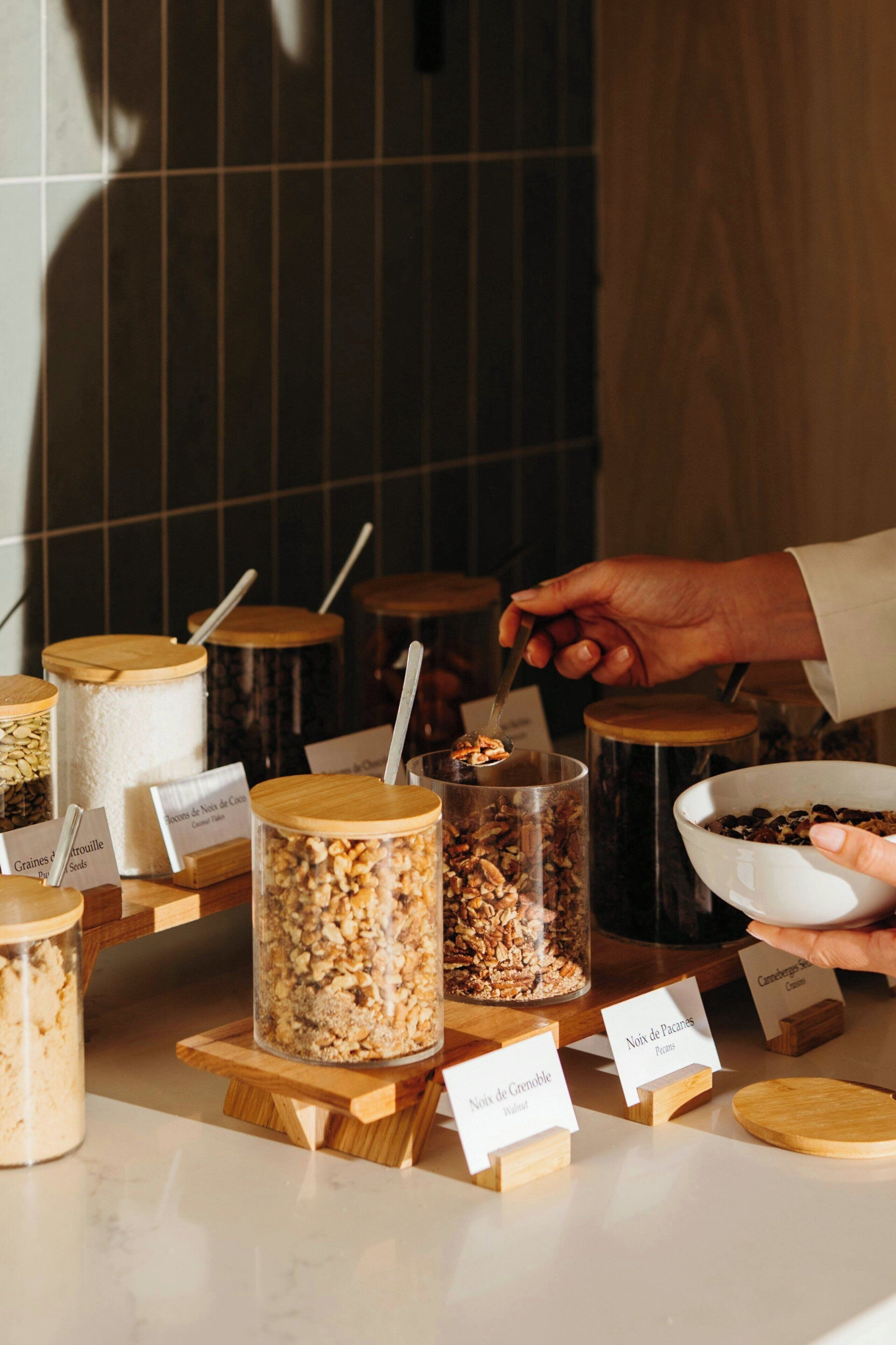 En række skåle med granola og frø i gennemsigtige beholdere med trälåg, hver mærket med ingrediensens navn.