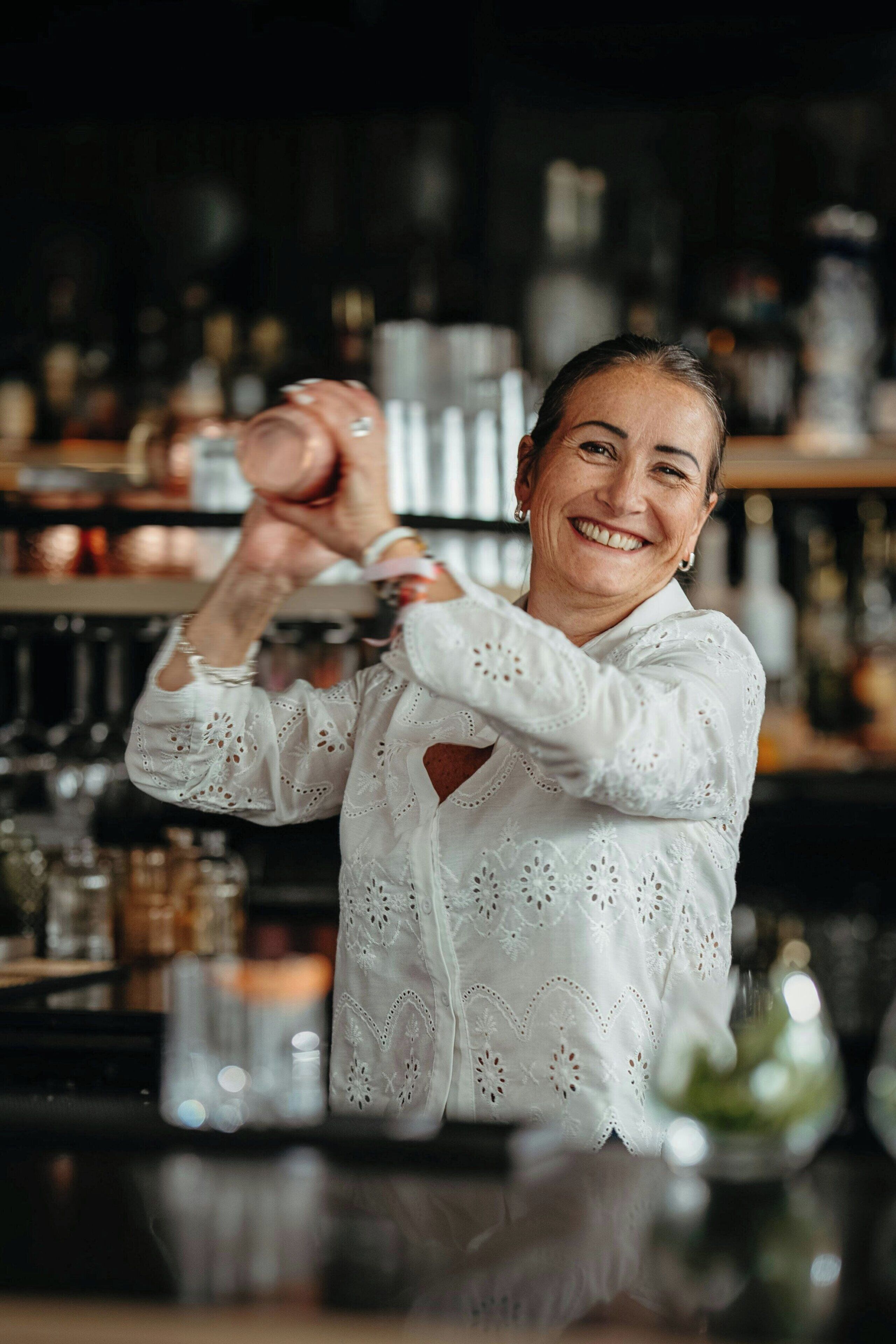 A person in a white shirt with lace details, standing in a bar setting with shelves of bottles and glasses in the background.