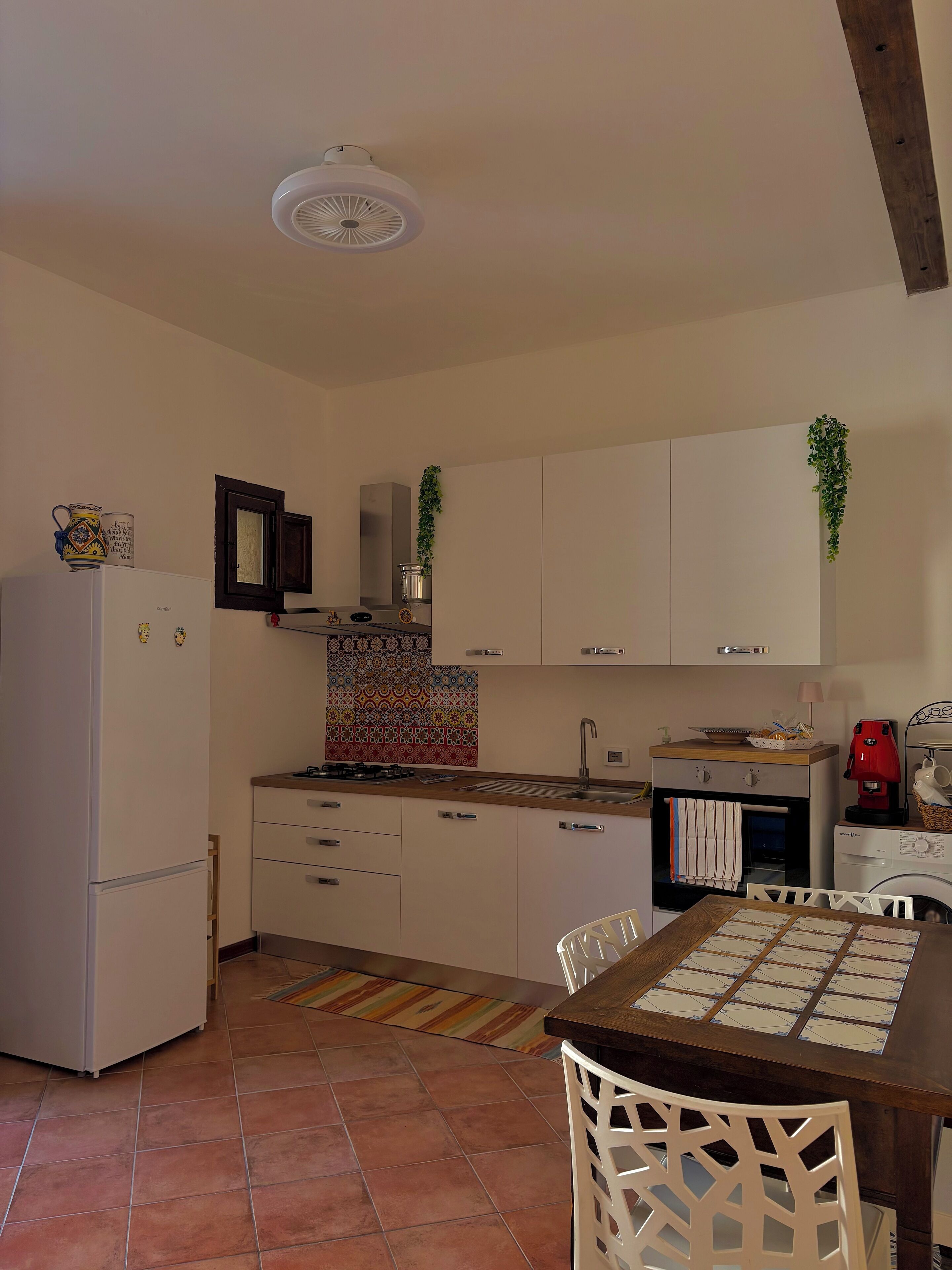Una cucina compatta con mobili bianchi, un frigorifero, un lavello e una macchina da caffè. Presente un pannello decorativo e una pianta pensile.