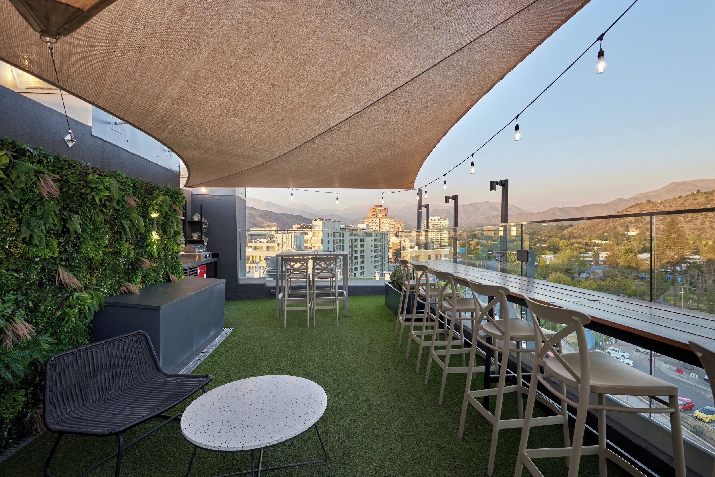 Un bar en la azotea con vistas a la ciudad, una pared verde y mesas al aire libre.