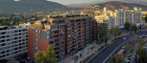 Un edificio de varios pisos con balcones, una carretera con vehículos y montañas al fondo.