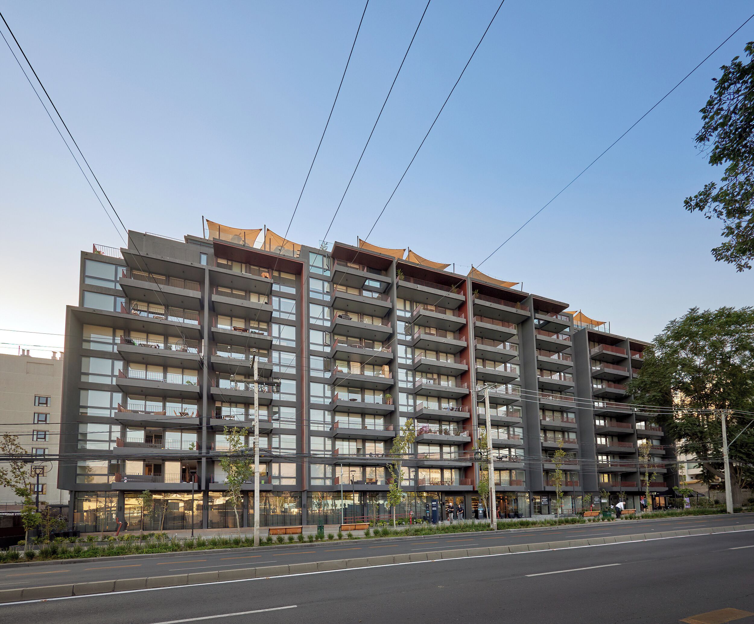 A multi-story residential building with balconies, a street in front, and a clear sky.