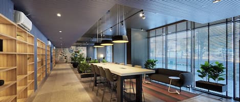 A modern conference room with a long table, chairs, and a sofa. There are potted plants and a large window with blinds.