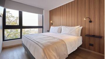 Un dormitorio con un ventanal grande, una cama con ropa de cama blanca, un panel de pared de madera y una lámpara de pared.