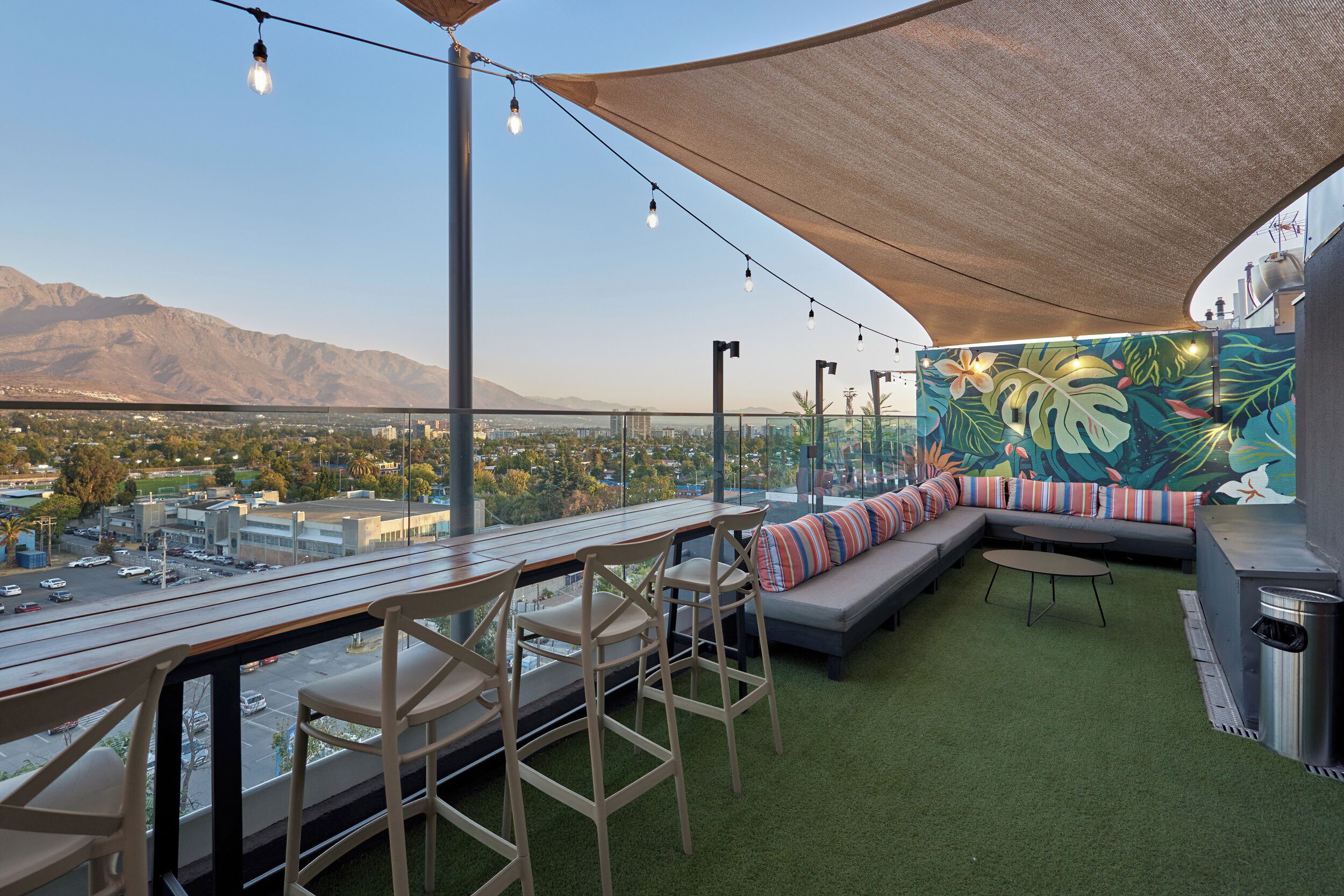 A rooftop terrace with a view of a city and mountains, a dining area with chairs and a table, and a seating area with a sofa and cushions.