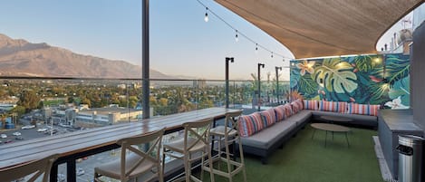 A rooftop terrace with a view of a city and mountains, a dining area with chairs and a table, and a seating area with a sofa and cushions.
