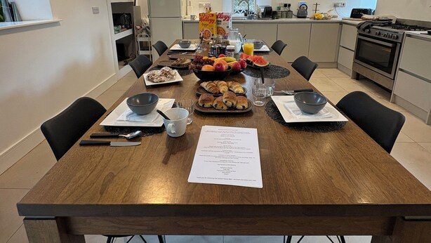 A modern kitchen with a wooden dining table set for breakfast, featuring croissants, fruits, and a bowl of cereal.