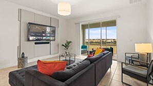 A modern living room with a large flat-screen TV, a dark sofa with colorful cushions, a small round coffee table, and a balcony with outdoor seating. - Stylish 3BR Condo Championsgate w Water Park (Davenport)