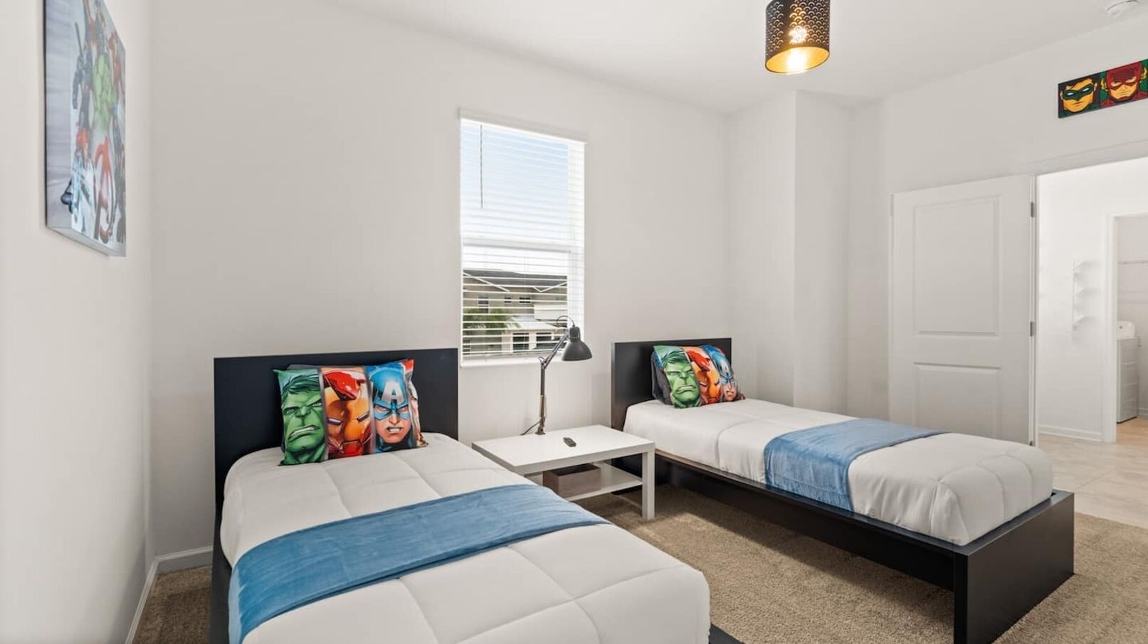 A modern bedroom with two beds, a small table, a lamp, and a window with blinds.