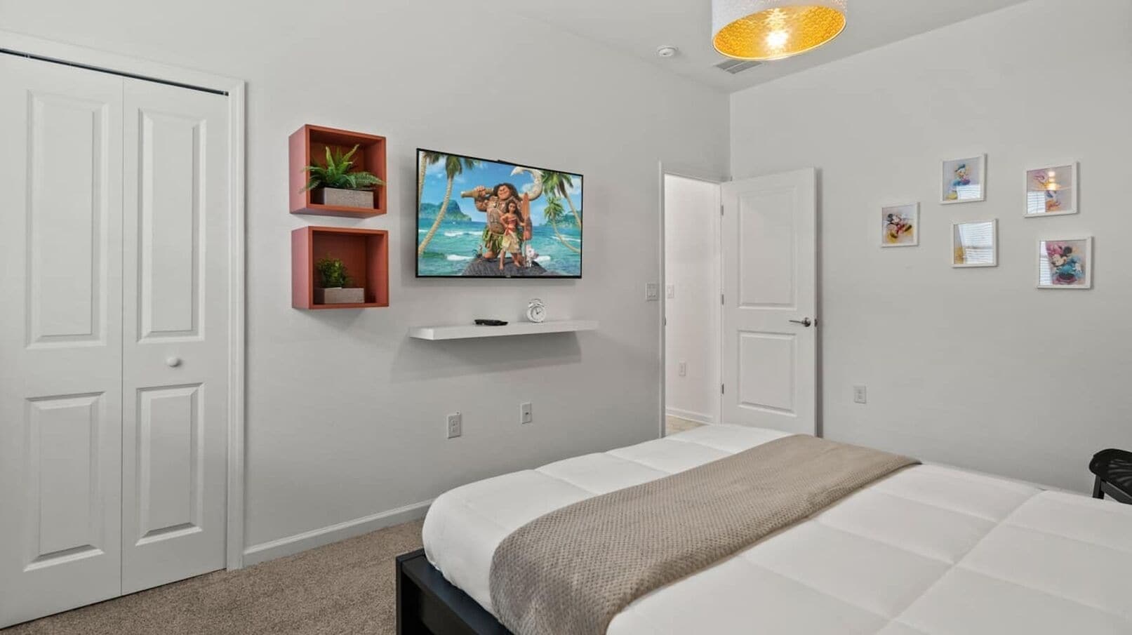 A bedroom with a bed, a TV mounted on the wall, a shelf with plants, and a door.