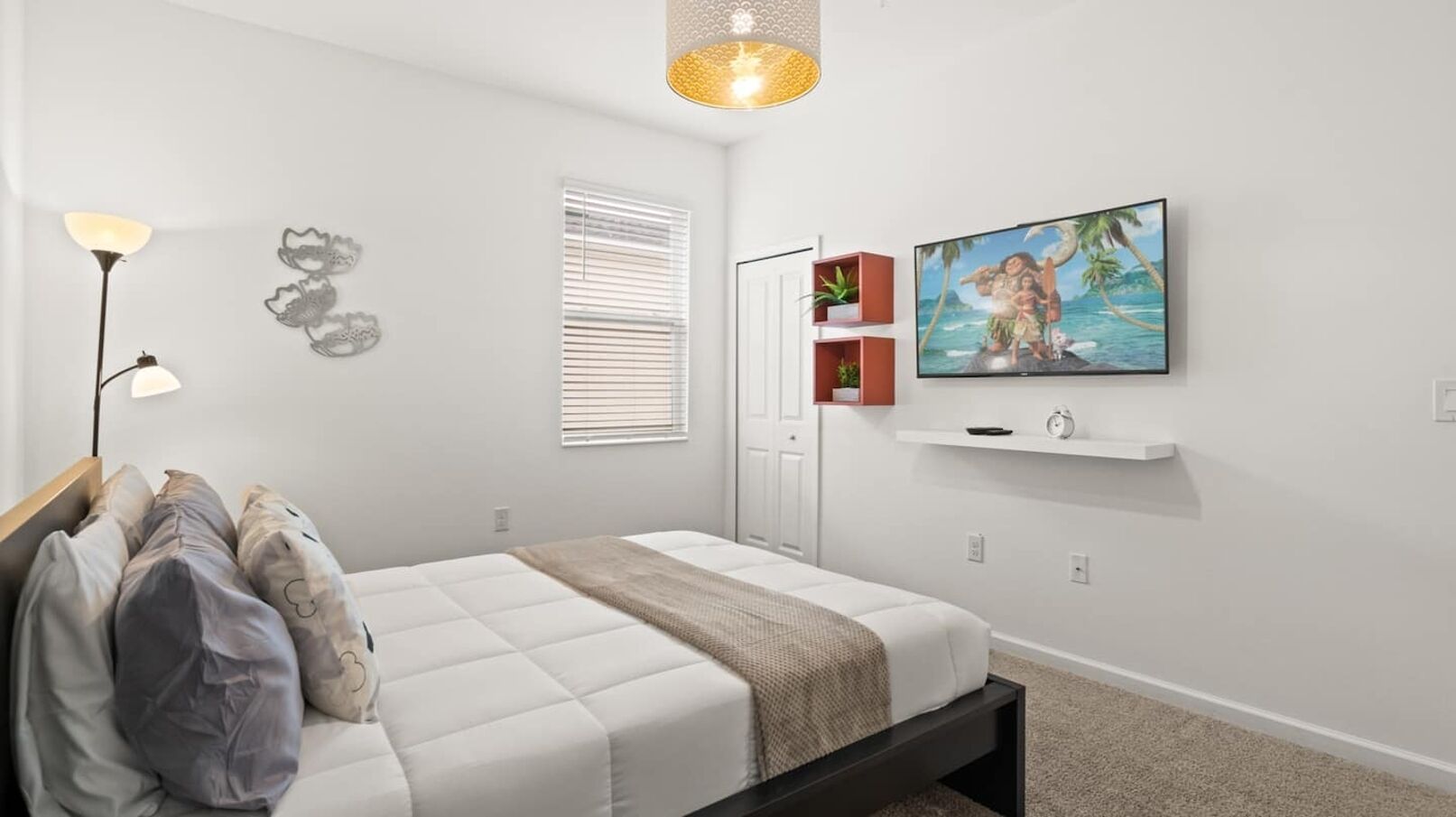 A bedroom with a bed, a bedside lamp, a wall-mounted TV, and a window with blinds.