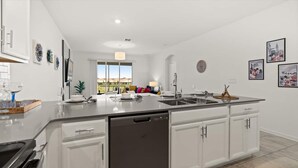 A modern kitchen with white cabinetry, a dishwasher, and a large island with a sink. There are decorative plates on the wall and a window with a view of a building. - Stylish 3BR Condo Championsgate w Water Park (Davenport)