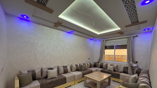 Un salon moderne avec un canapé d’angle, une table basse en bois et une vue sur l’extérieur grâce à une fenêtre agrémentée de rideaux.