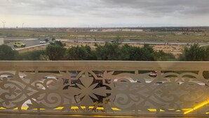 Une vue depuis un balcon orné de ferronnerie décorative, une route avec des véhicules et des bâtiments éloignés sous un ciel nuageux.