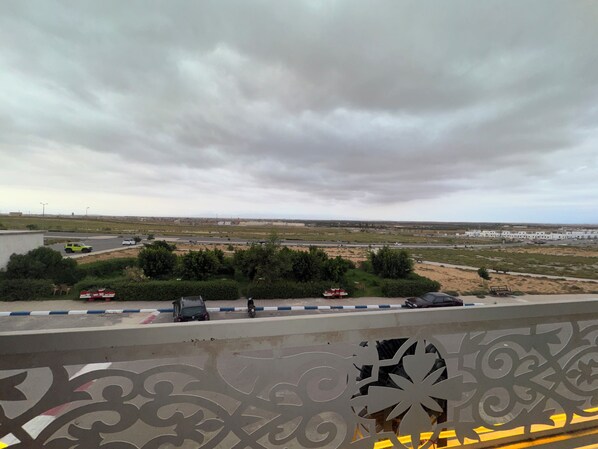 A view from a balcony overlooking a road with vehicles, a parking area, and distant buildings under a cloudy sky.