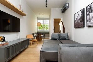 Ett modernt vardagsrum med en grå soffa, en platt-TV, en trähylla och en matplats med ett bord och stolar.