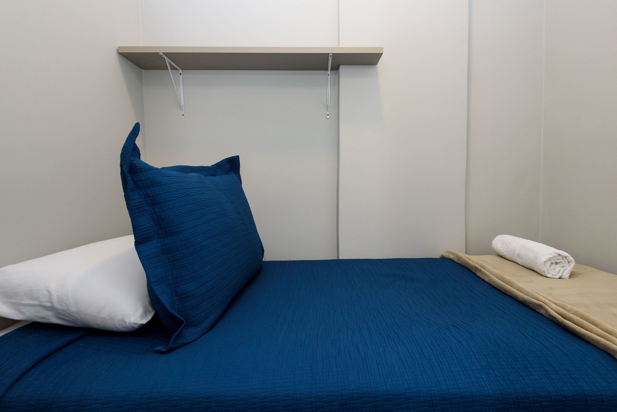 A single bed with a blue mattress, a white pillow, and a blue accent pillow. There is a beige shelf above the bed with a rolled white towel on it.